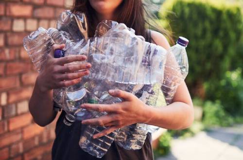 A woman stands outside by a brick wall with green bushes in the background, holding a collection of clear plastic bottle clutched in her arms