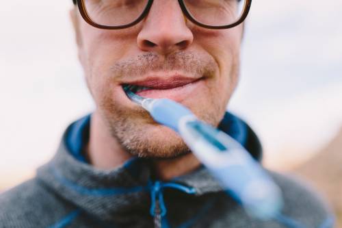 A blue toothbrush sticks out of a man's mouth towards the camera. The man wears a blue hooded top and black glasses, though  only the bottom of his glasses can be seen in the photo.