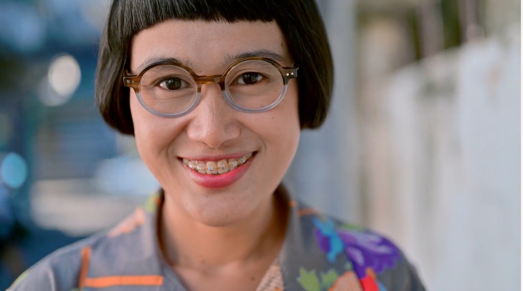 A women grins at the camera showing off the fixed braces on the top row of her teeth. She wears rounds glasses and has bobbed hair with a straight fringe across her forehead.