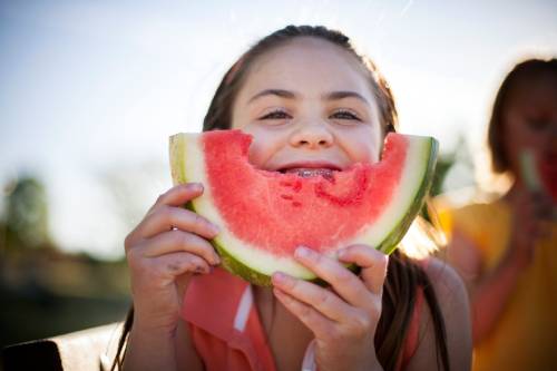 A young girl is eating a big slice of watermelon, holding it against her face so it looks like a smile. Through the hole made where she has been eating, you can see she is smiling and see the braces on her teeth.