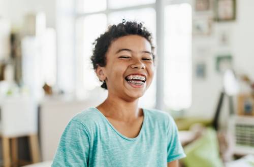 A smiling boy with short afro hair and a pale blue tee-shirt stands in a kitchen with his big open smile showing off the braces on his  teeth