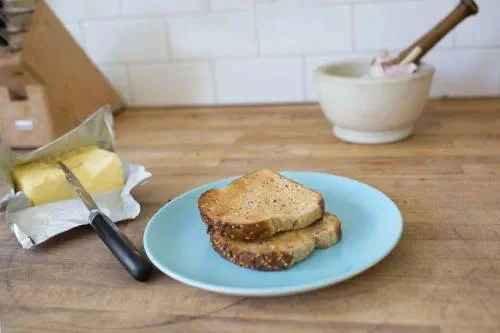 Two slices of toasted seedy brown bread sit on a pale blue plate with an open pack of butter and butter knife placed on the surface next to the plate